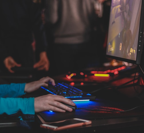 A person is engaged in a video game at a computer setup, using a keyboard and mouse. The scene is dimly lit, with the glow from the monitor reflecting off the hands of the player. Several other people are present in the background, indicating a social or competitive gaming environment.