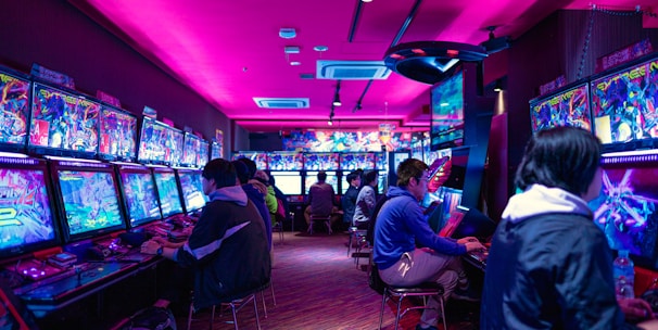 A vibrant arcade with rows of gaming machines adorned with colorful screens. Several people are immersed in playing games, seated on stools in a dimly lit room accented by an intense pink ceiling light. The atmosphere is lively and engaging.