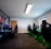 A well-lit gaming room features multiple gaming stations with high-backed chairs and monitors. The room has a minimalist setup with green and black color tones. Large posters adorn the walls, and the room appears clean and organized.