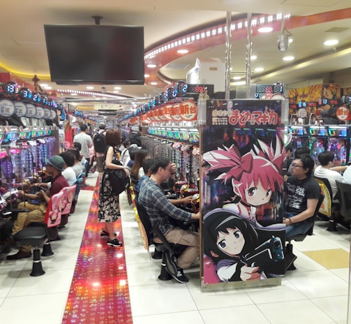A bustling arcade filled with people playing various gaming machines. Brightly colored displays and animated characters are prominent on posters and screens. The atmosphere is lively with patrons seated in rows, focusing on the games. Overhead signage and electronic scoreboards are visible, contributing to the busy and vibrant environment.