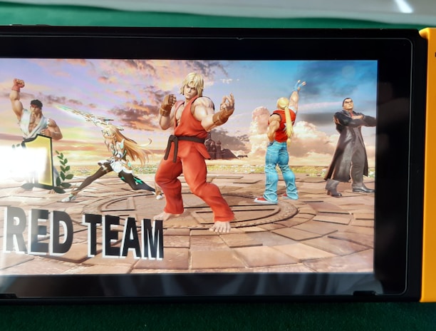 A handheld gaming console displays a screenshot of a fighting game featuring cartoonish characters in dynamic poses. The characters are standing in a virtual arena setting with a sky and cloud backdrop. The console is placed on a green surface.