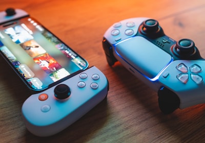 A gaming scenario featuring a smartphone with a game interface next to two controllers on a wooden surface. The smartphone displays vibrant images and icons, while the controllers feature a sleek, modern design with buttons and joysticks prominently visible. Warm lighting enhances the scene.