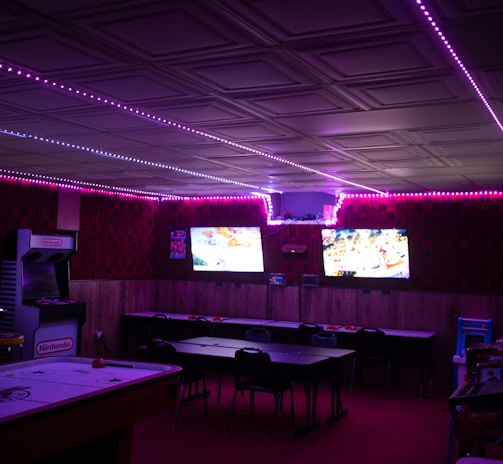 A dimly lit room with vibrant purple LED strip lights on the ceiling, featuring multiple gaming stations. There are two large screens showing video games, along with various arcade machines, including a Nintendo arcade cabinet. An air hockey table is positioned in the foreground.