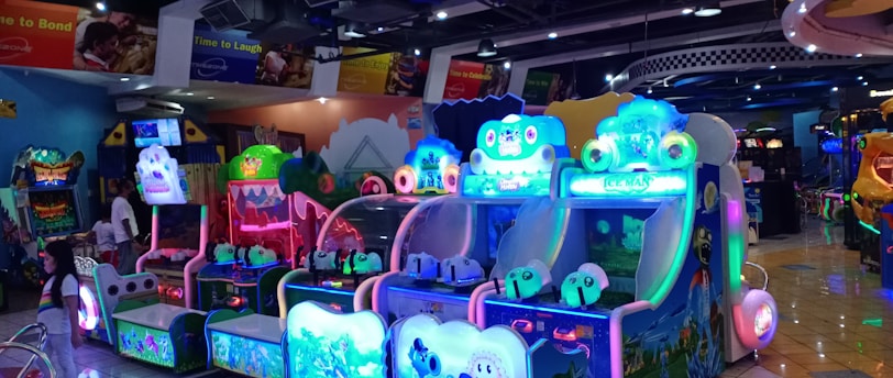 A brightly lit arcade filled with colorful gaming machines sits under ceiling lights. Neon lights illuminate various game stations, creating a playful and vibrant environment. A few red chairs are scattered around the reflective, tiled floor, and a person is observing one of the machines. The atmosphere is lively and cheerful, featuring games with cartoonish designs and engaging themes.
