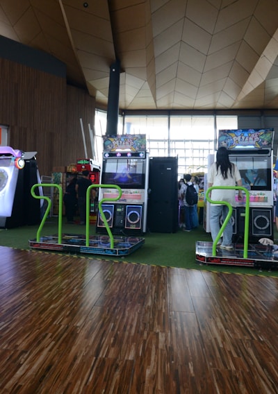 A vibrant arcade setting with several gaming machines lined up, including dance and rhythm game setups. People are engaged with the games, absorbed in their activities. The flooring is wooden, and the ceiling has a geometric design. Natural light filters through large windows, creating a lively atmosphere.