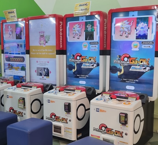 Several arcade gaming machines with multiple screens displaying Pokémon-themed content are positioned in a colorful arcade setting. There are people interacting with the machines; some are seated, and others are standing. The area features different gaming options, including a jumping game with bright, inviting graphics.